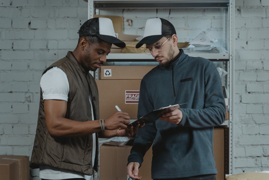 Two male couriers discussing and signing a clipboard in a warehouse setting, surrounded by packages.